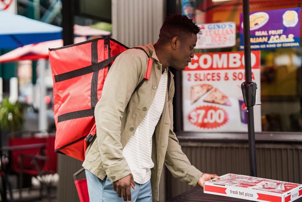 A pizza delivery person with a red backpack collects an order from a pizzeria.